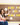 A brunette teacher in a white, sequined t-shirt with apple motif and blue flared jeans points to a sign in her classroom that reads "Kind people are my kinda people." The teacher is sitting on a table in her high school classroom.