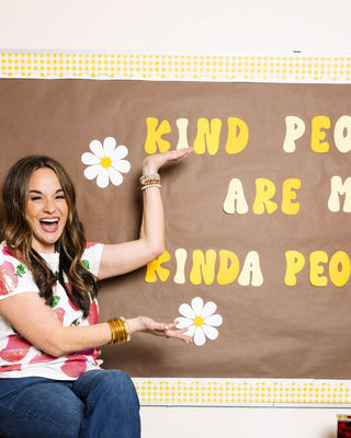 A brunette teacher in a white, sequined t-shirt with apple motif and blue flared jeans points to a sign in her classroom that reads "Kind people are my kinda people." The teacher is sitting on a table in her high school classroom.